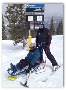 Photo of Cynthia Dietrich on a sled with hand skiis at the top of a mountain with an assistant behind her