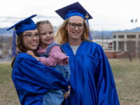 A woman with brown hair in a blue graduation cap and gown holds a young child in a pink shirt with overalls; a woman with blonde hair in a blue cap in gown stands on the right.