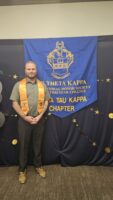 A man with blond hair and a gold stole poses in front of a blue banner that says Phi Theta Kappa.