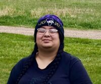 Trinity, wearing a hat, black shirt and jeans sits on a bench.outdoors.