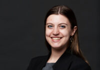 A woman in a gray top with a black cardigan poses for a headshot in front of a black background